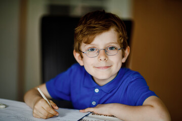 Portrait of little kid boy with glasses at home making homework, writing letters with colorful pens. Little child doing exercise, indoors. Elementary school and education, home schooling concept.