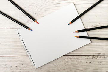 Notebook with white sheets and colored pencils for drawing on white wooden background
