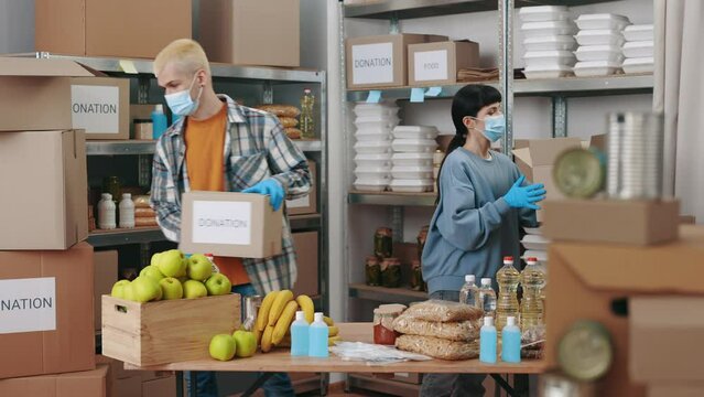 Charity workers in face masks and gloves standing together at storage and preparing boxes with food for donation. Volunteering, coronavirus and people concept.