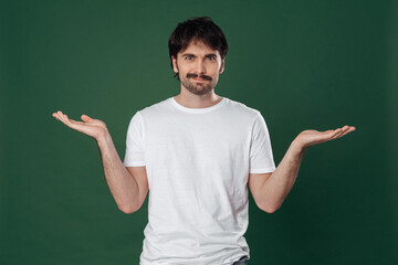 On a dark green background, a young man in a white T-shirt spread his arms to the sides and looks straight into the frame