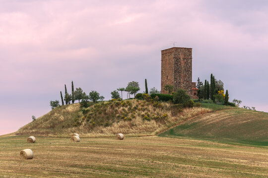 A Medieval magnificent Torre Tarugi and a rustic old house on a hill with bales of hay and trees around. Val d’Orcia, Italy