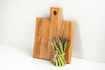 On a white background, a wooden board stands against the wall and a bunch of fresh green asparagus stands