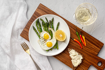 A white plate with scrambled eggs and asparagus stands on a wooden board. View from above. Nearby is a glass of white wine and toast with homemade cheese. 
