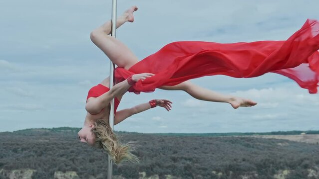 Young female pole dancer performs acrobatic tricks on pylon outdoors. Waving red cloth. Beautiful woman with slim body dancing. Sky and lake at background. 4K, UHD