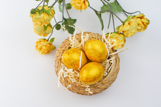 painted easter golden eggs in a wicker straw basket with yellow flowers on a white flat background, background for text