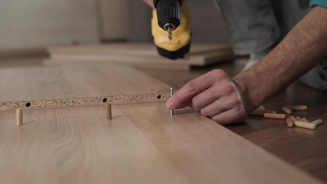 Caucasian boy puts screw into chipboard hole near wooden dowel. Male hands are twisting metal fastener with screwdriver, close-up. Dad and son assemble furniture, DIY repair.