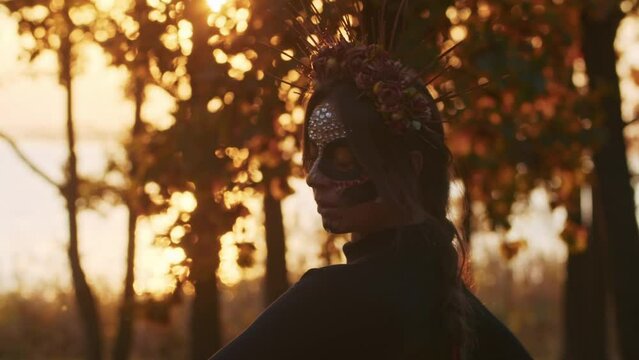 A young woman with Santa Muerte make-up dressed in a black dress of death walks against the backdrop of autumn leaves in the forest during sunset. Day of the Dead or Halloween concept.