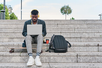 front view of an african american man sitting on a staircase working on his laptop. hipster man listening to music with yellow headphones.