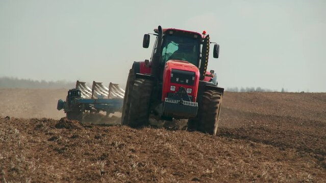 Modern tractor plows a field, moving for the camera. Front view of a red farm machine with a farmer picking up dust from the dry surface. Problem of soil erosion. Spring field work