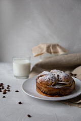 Composition with a bun on a white plate sprinkled with powdered sugar in the foreground, with a glass and a jar of milk in the background, scattered coffee beans on a light gray background 