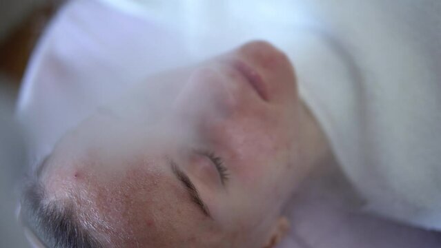 Close up of young woman with eyes closed having thermal steam treatment on problematic skin. Girl wearing white gown laying on couch in cosmetic salon
