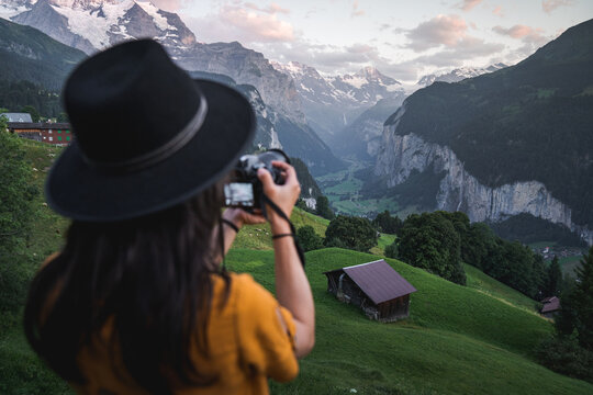 Young woman tourist taking photos with digital camera at famous touristic place in Wengen during sunset. Famous Instagram hiking place with green fields and snowy mountains over Lauterbrunnen Valley