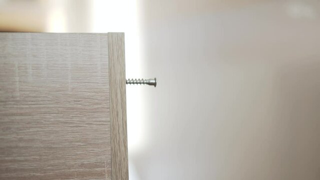 Man hand inserts and picks up galvanized euro confirm screw to connect two perpendicular furniture pieces made of chipboard, close-up.