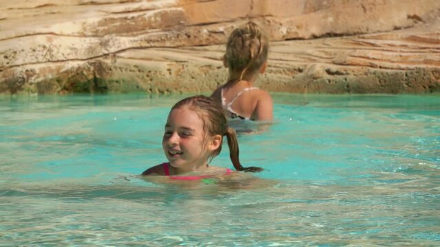 Two sibling girls are playing in swimming pool on background of artificial rock