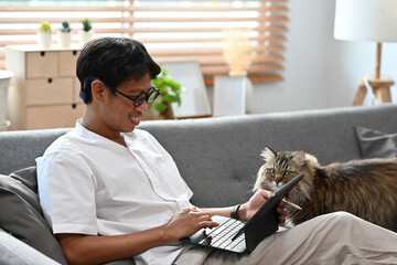 A portrait of a smiley Asian man working on a tablet at home and having a cat with him on the couch, for business, education, home and technology concept.