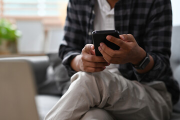 Cropped view Asian man sitting on a couch in a living room using a smart phone and a laptop, for business, communication, home and technology concept.