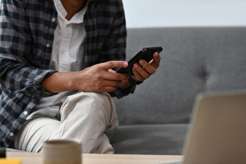 Cropped view Asian man sitting on a couch in a living room using a smart phone and a laptop, for business, communication, home and technology concept.