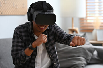 A portrait of a man sitting on a couch in a living room, wearing a virtual reality technology goggle and acting fighting, for future, game and technology concept.