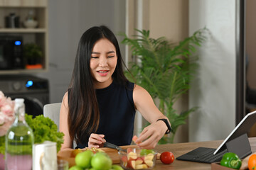 Obraz na płótnie Canvas A portrait of a young pretty Asian woman chopping fruits and preparing a meal on a wooden cut board, for food, health and cooking concept.
