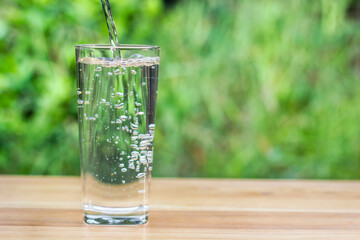 Pour water into glass on wooden table outdoors and green background.