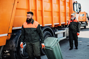 Senior and young garbage men working together on emptying dustbins for trash removal..
