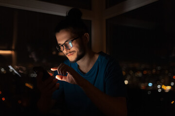 Night portrait of young man touching screen of smartphone. Window background.