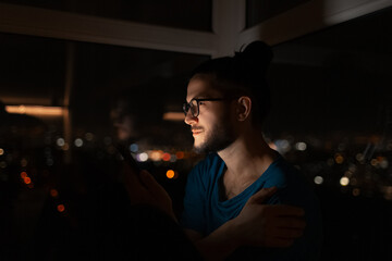 Night side portrait of young man holding smartphone near window background.