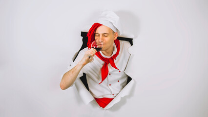 Young handsome man dressed as chef with glass of wine. Male cook drinking alcoholic beverage and sticking out of hole of white background.