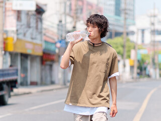 Portrait of handsome Chinese young man with curly black hair in brown T-shirt and pants drinking water while walking on Shanghai old town street, front view of cool young man.