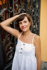 Fototapeta premium Beautiful brunette young woman in a beautiful white summer dress walks and poses in the summer in the city.