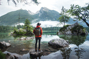 Rear View Of Man with backpack, hat and orange sofshell jacket Looking At Lake Against Mountains,...