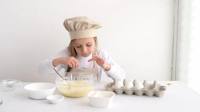 Small cute girl in special clothes and chef cap tastes and eats sugar from spoon. Kid enjoy and learning to cook. Child  lick fingers. On white table stand eggs and in bowls sugar, salt and soda