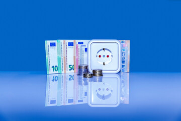 Euro banknotes and coins stacked. White European plug socket on blue background. Concept of saving electricity at home. Increasing of electricity cost, expensive energy, rise in electricity prices