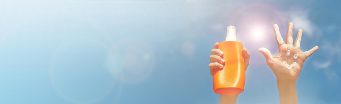 Banner of positive symbol drawing by sunscreen on open hand, another hand holding a bottle of suntan lotion. Two hands on blue sky, clouds, beams of sun background.