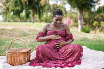 Happy pregnant african woman sitting at city park and touching her belly