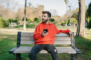 Young man sitting on a park bench and looking away with phone. Holding smartphone, thinking over text message for online chat