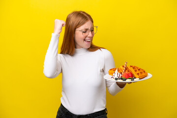 Young redhead woman holding waffles isolated on yellow background celebrating a victory