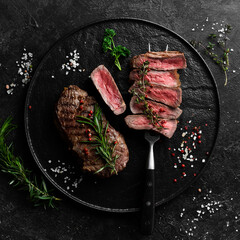 Grilled ribeye beef steak. Steak on a fork on a black plate. Top view. On a black stone background.