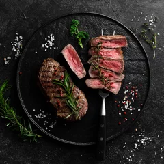 Foto op Canvas Grilled ribeye beef steak. Steak on a fork on a black plate. Top view. On a black stone background. © Yaruniv-Studio