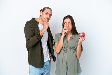 Young caucasian couple isolated on white background pregnant and holding an apple while thinking