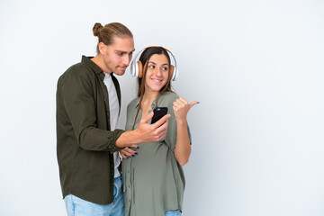 Young caucasian couple isolated on white background pregnant and listening music while pointing side