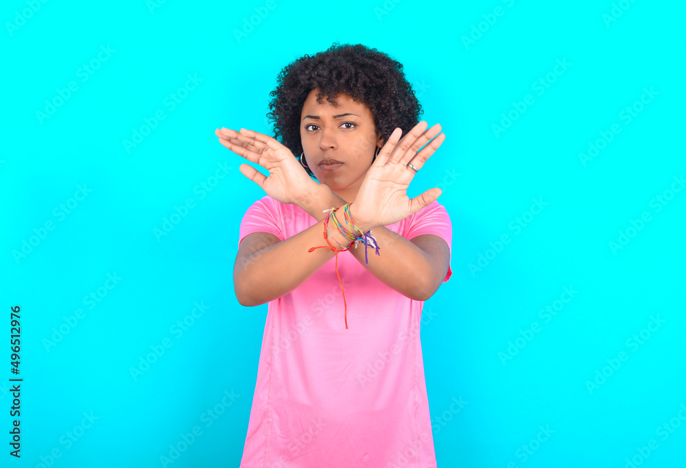 Wall mural young girl with afro hairstyle wearing pink t-shirt over blue background has rejection expression cr