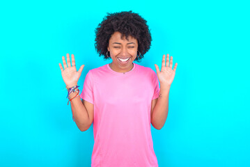 Emotive young girl with afro hairstyle wearing pink T-shirt over blue background laughs loudly, hears funny joke or story, raises palms with satisfaction, being overjoyed amused by friend