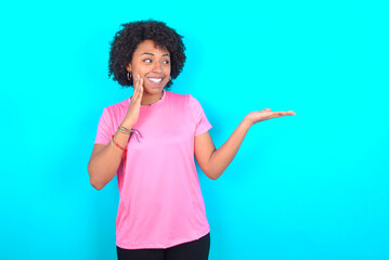 Funny young girl with afro hairstyle wearing pink T-shirt over blue background holding open palm new product. I wanna buy it!