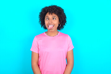 young girl with afro hairstyle wearing pink T-shirt over blue background showing grimace face crossing eyes and showing tongue. Being funny and crazy