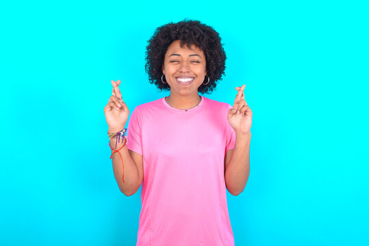 young girl with afro hairstyle wearing pink T-shirt over blue background has big hope, crosses fingers, believes in good fortune, smiles broadly. People and wish concept