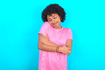 young girl with afro hairstyle wearing pink T-shirt over blue background Hugging oneself happy and positive, smiling confident. Self love and self care