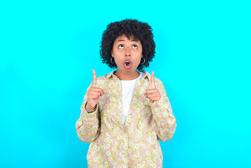 young girl with afro hairstyle wearing floral shirt over blue background amazed and surprised looking up and pointing with fingers and raised arms.