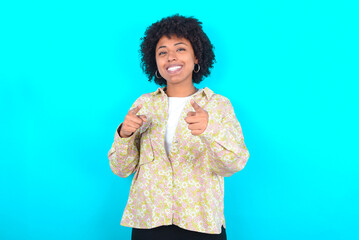 young girl with afro hairstyle wearing floral shirt over blue background pointing fingers to camera with happy and funny face. Good energy and vibes.