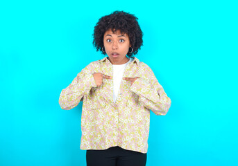 Embarrassed young girl with afro hairstyle wearing floral shirt indicates at herself with puzzled expression, being shocked to be chosen to participate in competition, hesitates about something